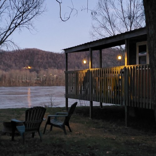 A peaceful riverside scene at dusk, featuring a wooden deck with chairs and soft lights, surrounded by trees and hills.