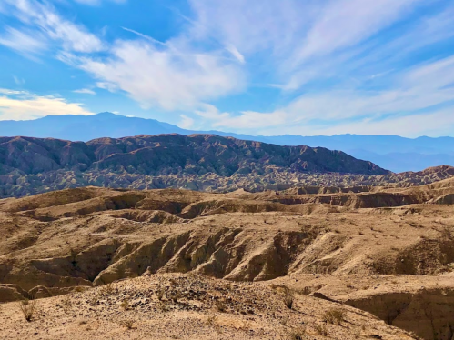A vast desert landscape with rugged hills under a blue sky and wispy clouds in the background.
