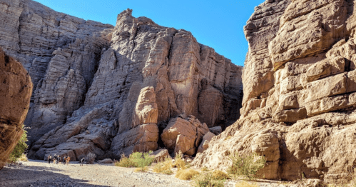 A rocky canyon with towering cliffs and a clear blue sky, featuring a small group of hikers in the distance.