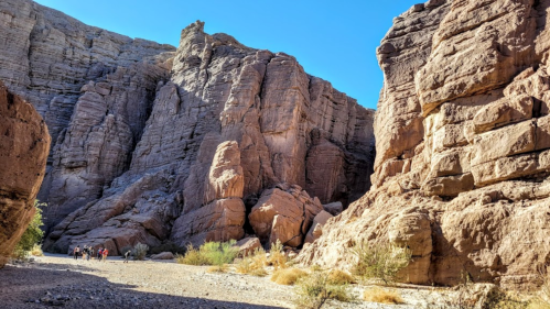 A rocky canyon with towering cliffs and a clear blue sky, featuring small figures walking along a dry riverbed.