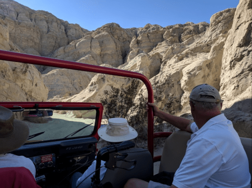 Two people in a vehicle view rocky cliffs in a desert landscape under a clear blue sky.