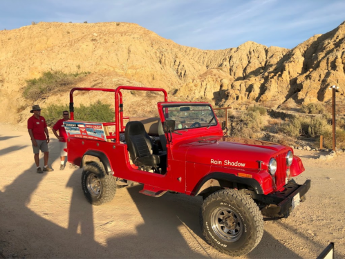 A red jeep parked on a dirt path with rocky hills in the background; a person stands nearby.