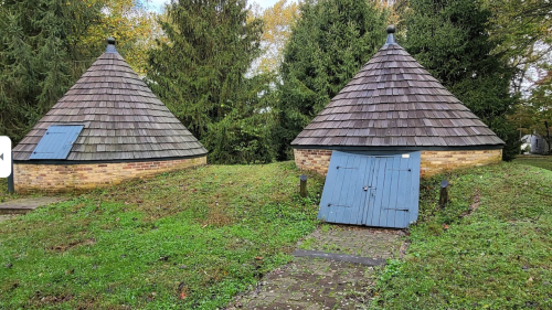 Two conical-roofed structures with blue doors, surrounded by greenery and trees, on a stone pathway.