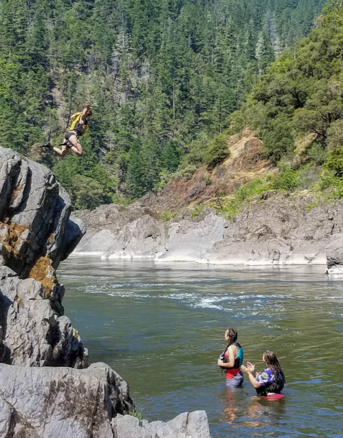 A person jumps off a rock into a river while two others watch from the water's edge in a scenic, wooded area.