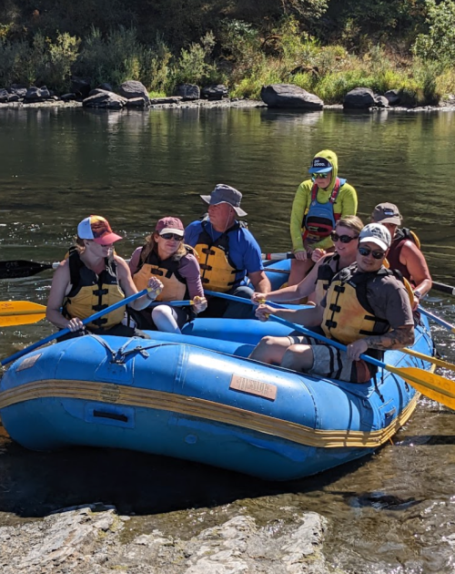 A group of eight people in life jackets paddling a blue raft on a calm river surrounded by greenery.