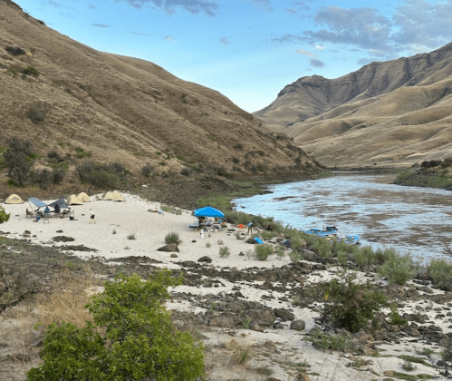 A sandy riverside campsite with tents and a blue canopy, surrounded by dry hills and a flowing river.