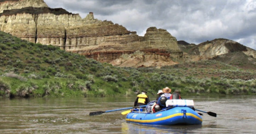 A group of people in a blue raft paddles down a river surrounded by rocky cliffs and green vegetation under a cloudy sky.