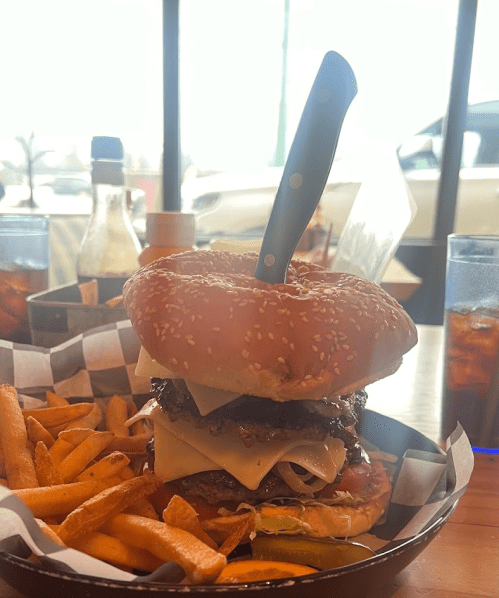 A towering burger with a knife in it, served with fries, on a table with drinks in the background.