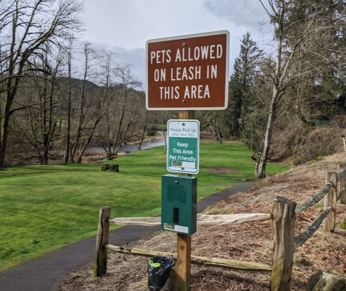 Sign indicating pets are allowed on leash, with a reminder to pick up after them, near a grassy area by a river.