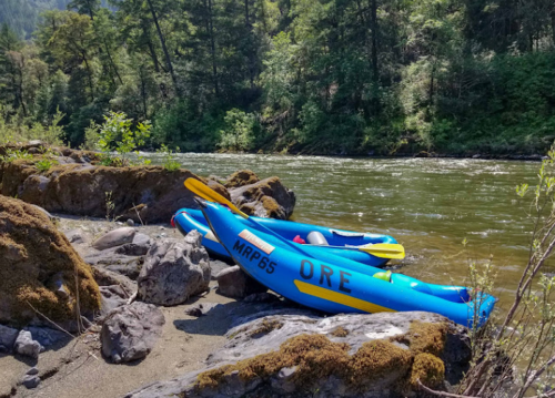 Two colorful kayaks resting on rocky shore by a river, surrounded by lush greenery and trees.