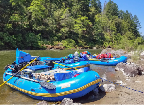 Two blue rafts are moored on a rocky riverbank, surrounded by lush green trees under a clear blue sky.