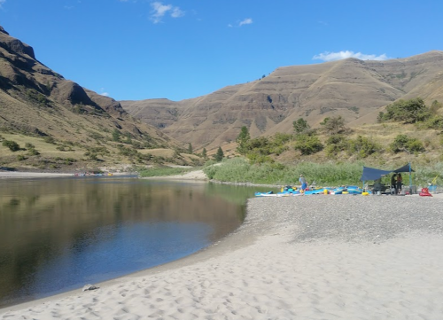 A serene riverbank scene with sandy shores, mountains in the background, and a campsite with kayaks nearby.