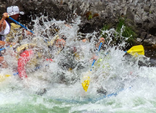 A group of people white-water rafting, splashing through turbulent water with paddles raised.