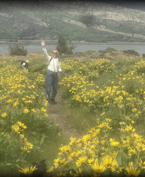 A person in a white shirt and suspenders waves while walking through a field of yellow flowers with a dog nearby.