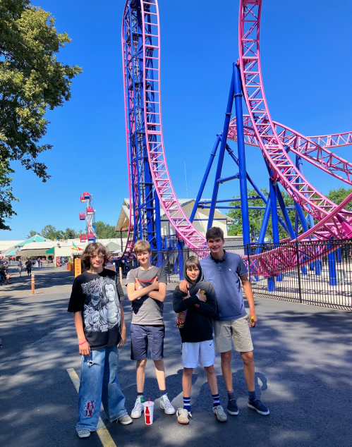 Four boys stand in front of a colorful roller coaster at an amusement park on a sunny day.