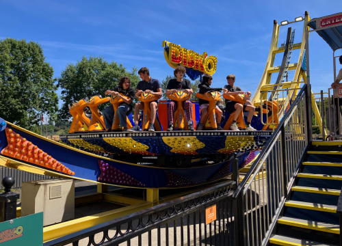 A group of four people sits on a colorful amusement park ride, ready for a thrilling experience under a clear blue sky.