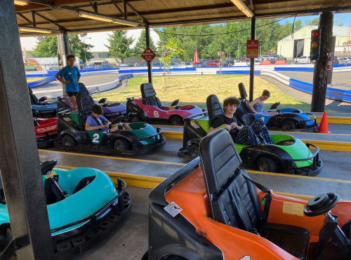 A go-kart track with several colorful karts and kids preparing to race under a shaded area.