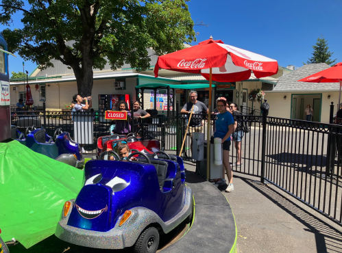 A colorful amusement park ride with people standing nearby, shaded by red umbrellas on a sunny day.