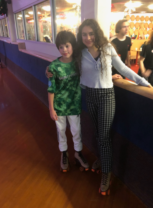 A boy in a green jersey and a girl in a white top and checkered pants pose together on roller skates at a rink.