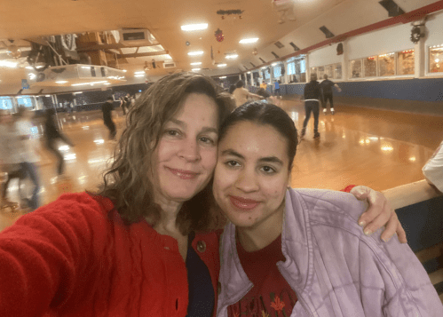 Two women smile for a selfie at a roller skating rink, with skaters in the background.