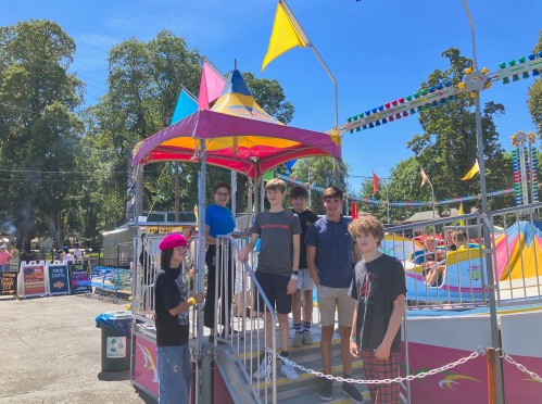 A group of five teenagers stands on a carnival ride platform, colorful flags and attractions in the background.