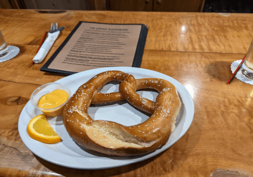 A large pretzel with salt, served on a plate with a side of cheese sauce and a slice of orange, on a wooden table.