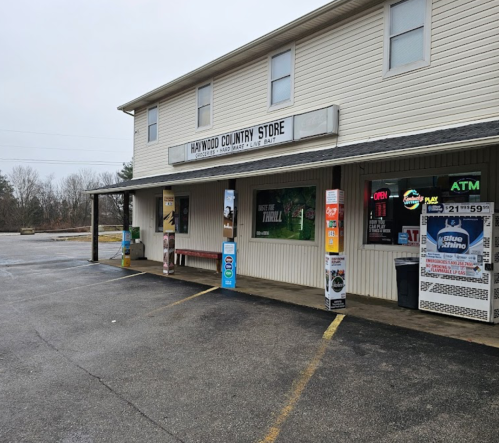 A small country store with a covered porch, featuring signs for open hours, an ATM, and various advertisements.