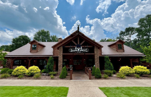 A rustic log cabin restaurant with a sign, surrounded by greenery and a blue sky with fluffy clouds.