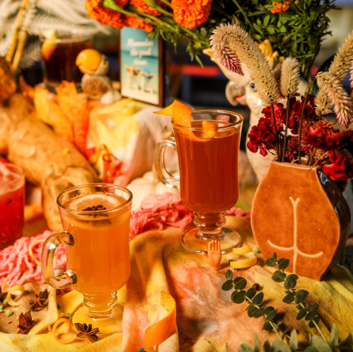 Two decorative glasses of colorful cocktails sit on a vibrant table, surrounded by flowers and festive decor.