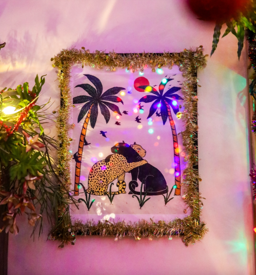 A colorful artwork of two animals under palm trees, framed with tinsel and surrounded by festive lights.