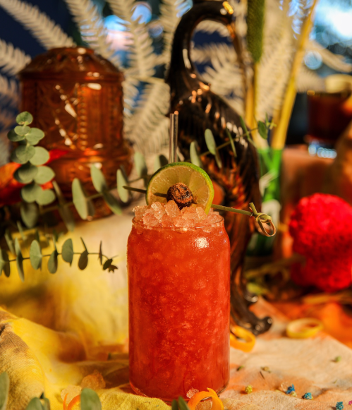 A vibrant cocktail garnished with a lime slice and a cherry, surrounded by decorative foliage and colorful accents.