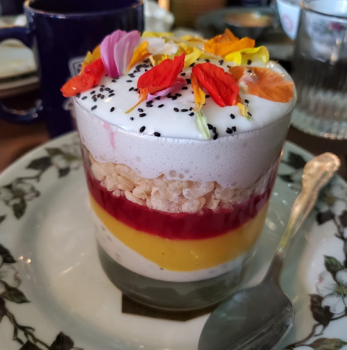 A colorful dessert in a glass, layered with rice crisps, fruit puree, cream, and topped with edible flowers.