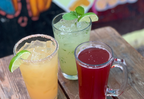 Three colorful drinks on a wooden table: a yellow cocktail with a salted rim, a green mint drink, and a red beverage.