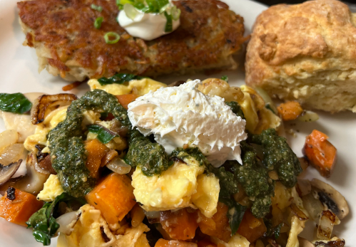 A plate featuring scrambled eggs with vegetables, pesto, a dollop of cream, a hash brown, and a biscuit.
