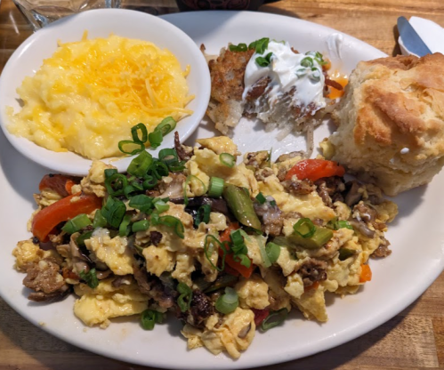 A plate of scrambled eggs with vegetables, a biscuit, cheesy grits, and a side of potatoes topped with sour cream.