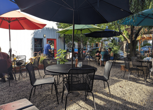 Outdoor seating area with tables and umbrellas in a cozy garden setting, featuring people enjoying their time.