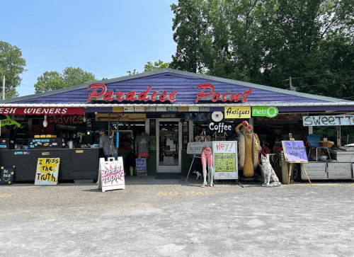 A colorful storefront of "Paradise Point" featuring signs for coffee, antiques, and fresh wieners, surrounded by greenery.