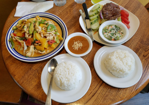 A wooden table with two plates of rice, a colorful vegetable dish, a side of sauce, and a plate of grilled meat with salad.