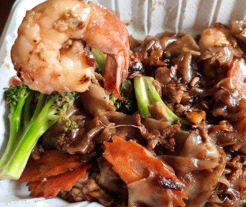 A close-up of a takeout container filled with shrimp, broccoli, and dark sauce over flat noodles.