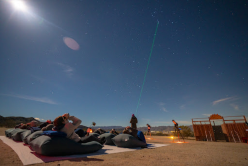 People lying on blankets under a starry sky, with a laser pointing towards the stars and telescopes set up nearby.