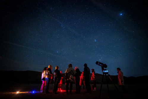 A group of people stargazing under a starry night sky, with a telescope and red lights illuminating the scene.