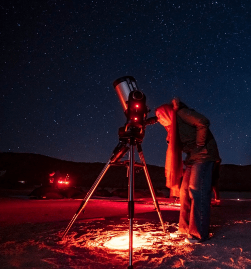 A person peers through a telescope under a starry night sky, with red lights illuminating the scene.