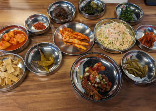A variety of small metal bowls filled with colorful side dishes, including pickled vegetables and salads, on a wooden table.