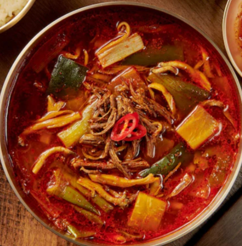A bowl of spicy beef stew with vegetables, garnished with red chili slices, served on a wooden table.