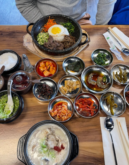 A table set with a hot stone bowl of bibimbap, a bowl of soup, and various side dishes in metal bowls.
