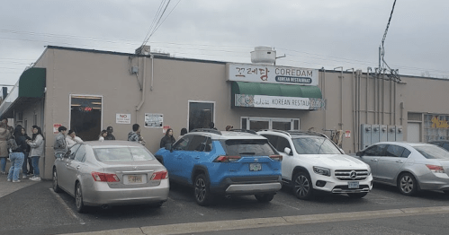 A busy Korean restaurant with a line of people outside and parked cars in front on a cloudy day.