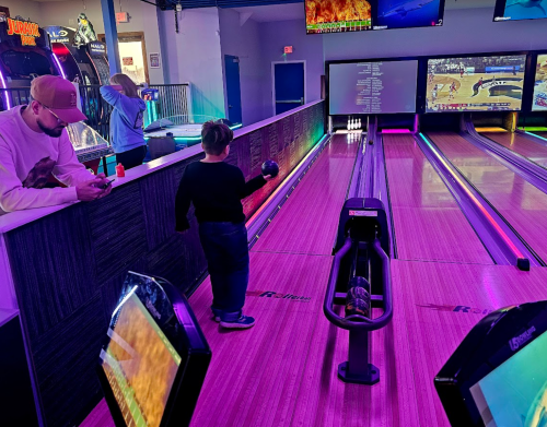 A child prepares to bowl in a colorful bowling alley, while an adult checks their phone nearby. Screens display scores.