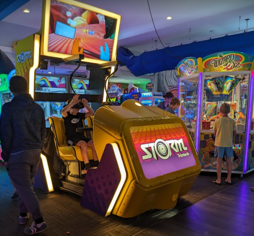 A vibrant arcade scene featuring a "STORM" ride and a colorful game cabinet, with kids enjoying the games.