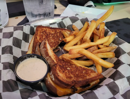 A basket with a grilled cheese sandwich, crispy fries, and a small cup of dipping sauce on a checkered paper.
