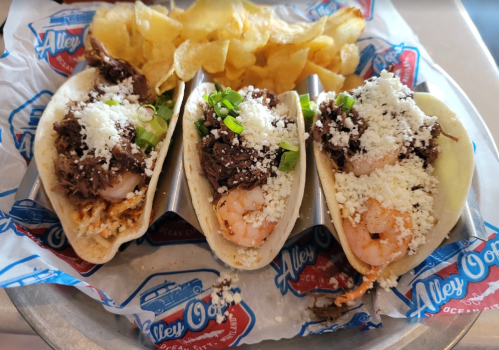 Three tacos filled with shrimp and beef, topped with cheese and green onions, served with potato chips on the side.